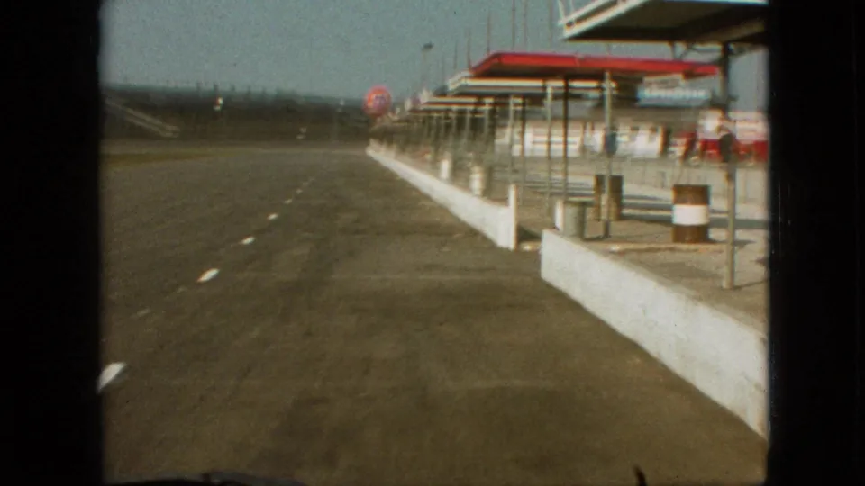1968 Branson, Missouri pit-area view from a moving vehicle, showing empty asphalt lanes, a low wall, service buildings, barrels, and a distant visible brand mark.