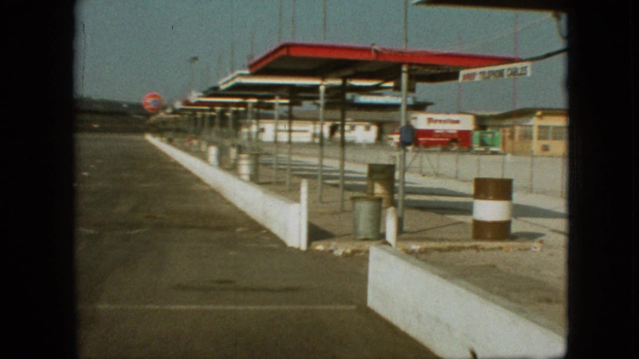 1968 Branson, Missouri home-movie frame showing a service station forecourt with a long white barrier, red-and-white canopy, Firestone truck, metal bins, and distant 76 sign.