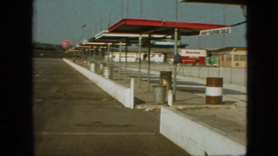 1968 Branson, Missouri home-movie frame showing a service station forecourt with a long white barrier, red-and-white canopy, Firestone truck, metal bins, and distant 76 sign.