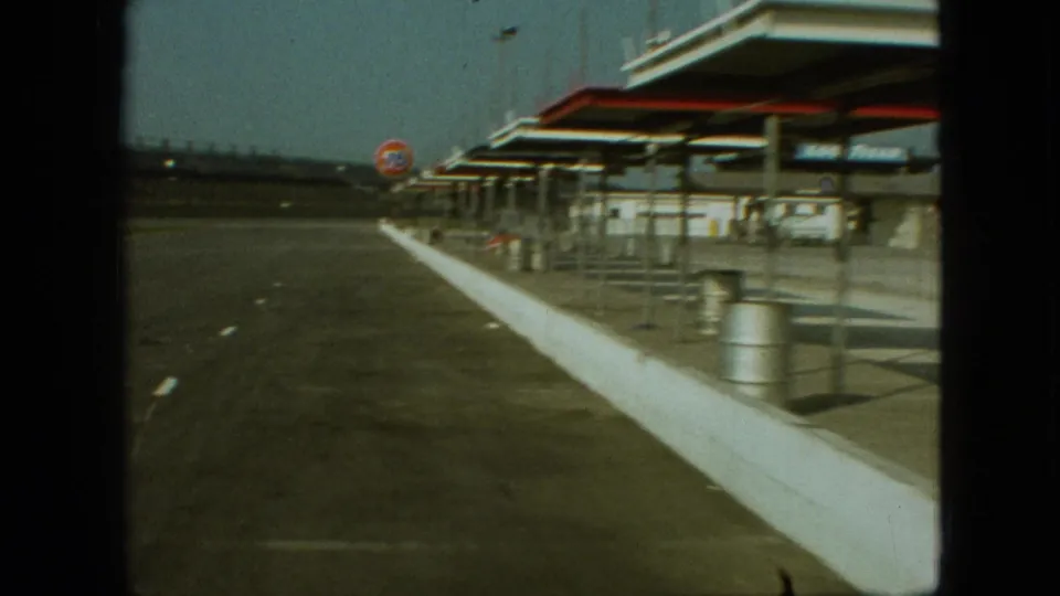1968 Branson, Missouri frame from a moving home-movie shot beside a gas station, showing the paved lane, white barrier, canopy posts, trash cans, and a circular 76 sign.