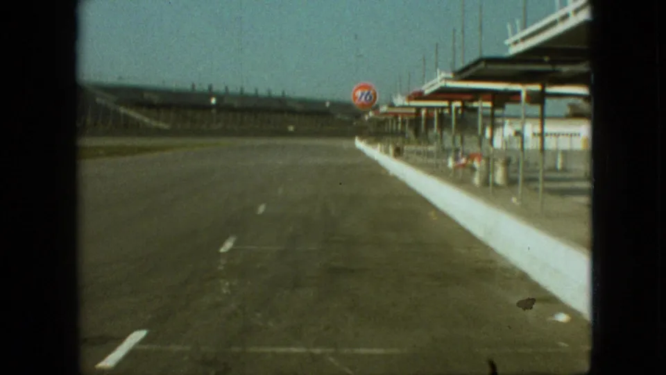 1968 Branson, Missouri home-movie frame showing the roadway alongside the station, with a white barrier, canopy structures, open pavement, distant hills, and a visible 76 sign.
