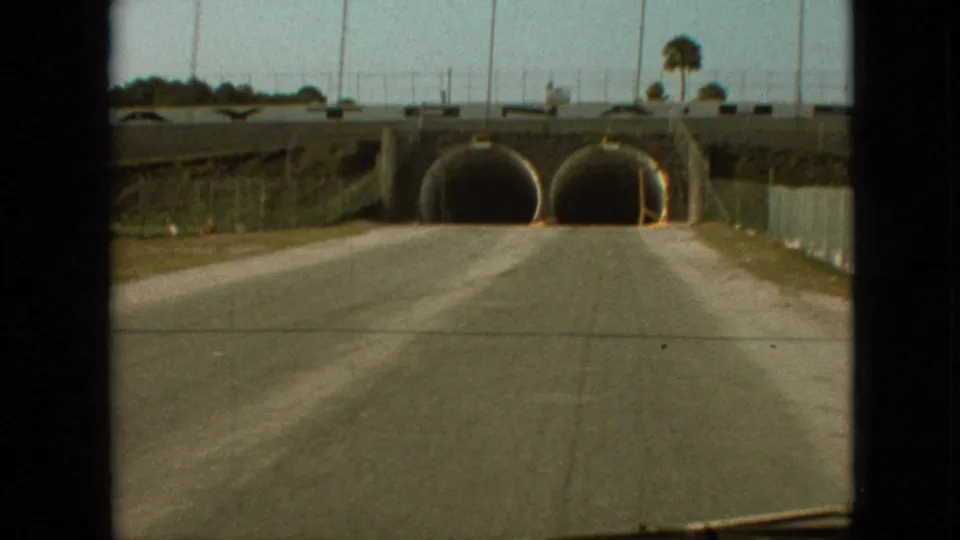 Branson, Missouri 1968 frame showing the car nearly at the twin tunnel entrances beneath an overpass, with open roadway, fencing, roadside poles, and wiper visible.