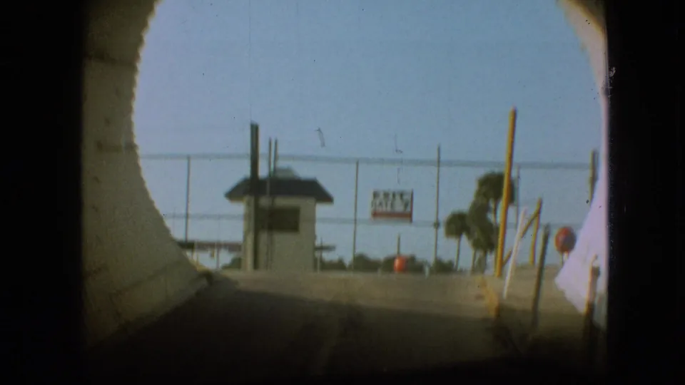 1968 Branson car POV emerging from the tunnel toward a chain-link fenced gate area with a small booth, signs, posts, and trees beyond.