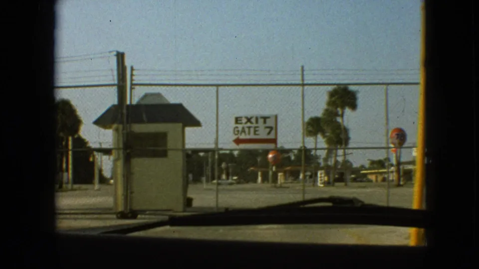 1968 8mm Branson car POV facing a chain-link gate, small booth, and white EXIT GATE 7 sign with a red arrow under a clear sky.
