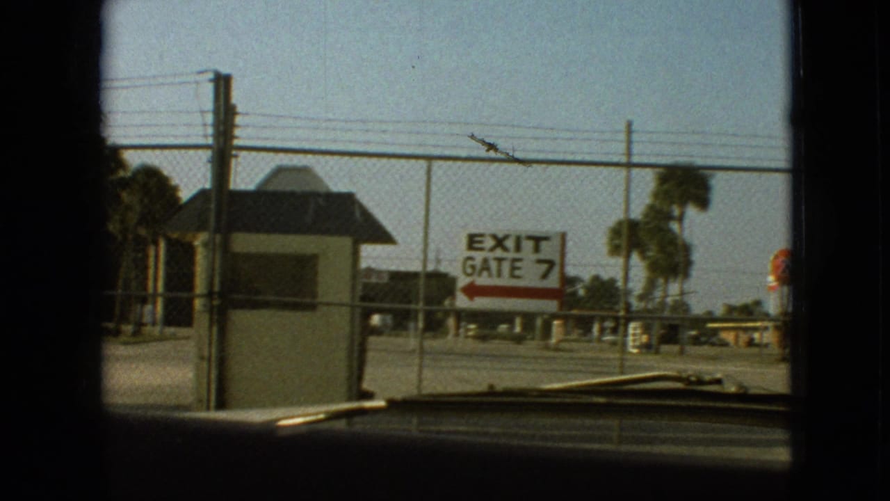 1968 8mm home-movie frame showing an EXIT GATE 7 sign, small guardhouse, fencing, palm-like trees, and a car windshield edge, with the exact facility not identified from the frame alone.