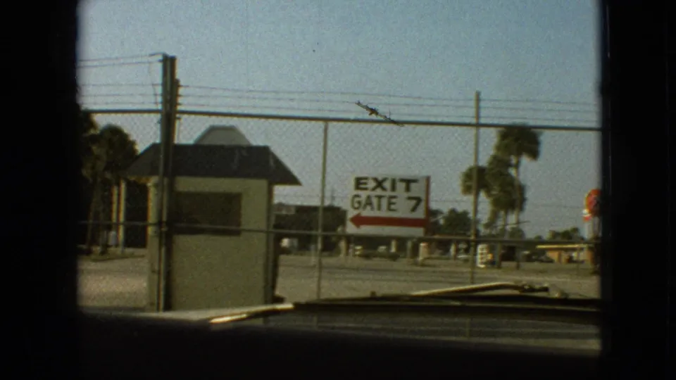 1968 8mm home-movie frame showing an EXIT GATE 7 sign, small guardhouse, fencing, palm-like trees, and a car windshield edge, with the exact facility not identified from the frame alone.