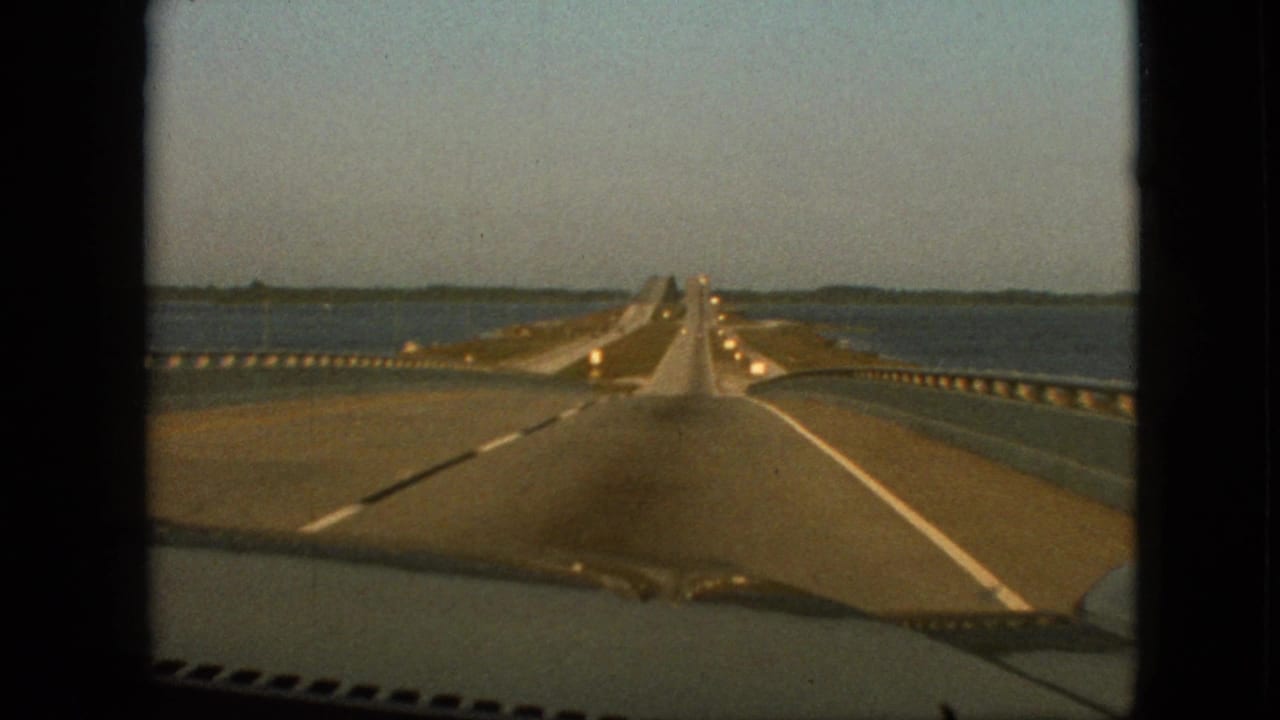 1968 8mm home-movie frame from inside a car looking down a long straight bridge over calm water, with vehicles ahead, guardrails, and a distant shoreline under clear sky.