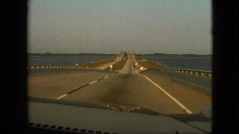 1968 8mm home-movie frame from inside a car looking down a long straight bridge over calm water, with vehicles ahead, guardrails, and a distant shoreline under clear sky.