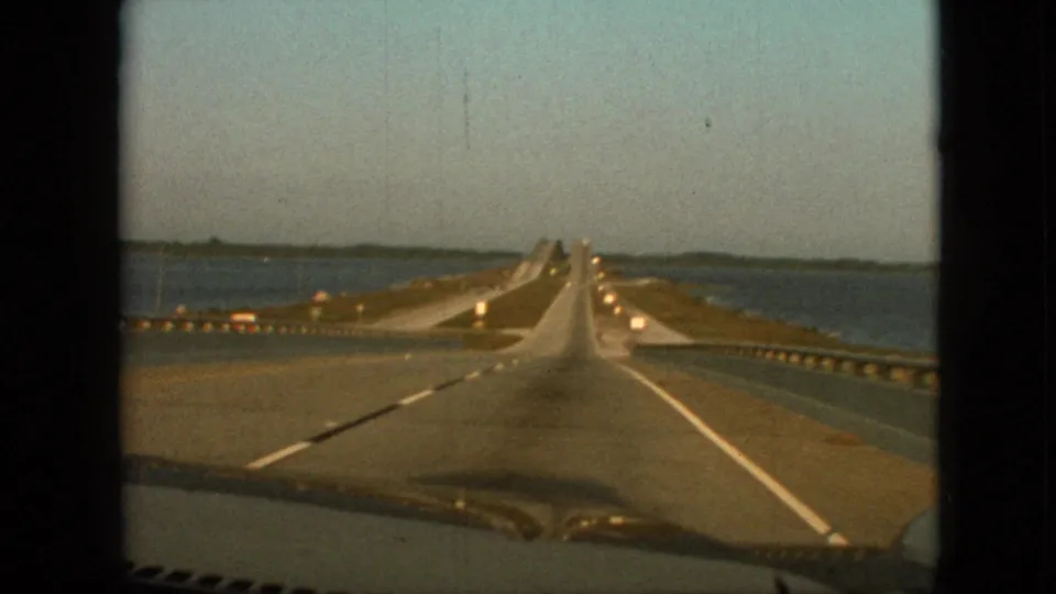 1968 8mm home-movie frame showing a forward car-window view of a bridge approach or span over water, with painted lane lines, guardrails, low shoreline, and dashboard edge visible.