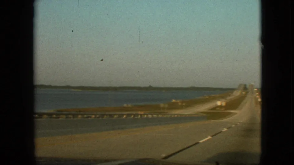 1968 8mm home-movie frame showing a road curving beside open water, with a guardrail, grassy embankment, distant shoreline, and the car hood or dashboard at the lower edge.