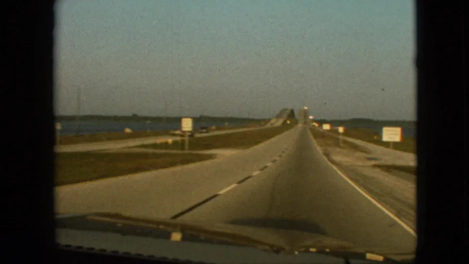Forward-facing 1968 home-movie frame from a moving car on a Missouri highway approaching a raised bridge or causeway near open water.