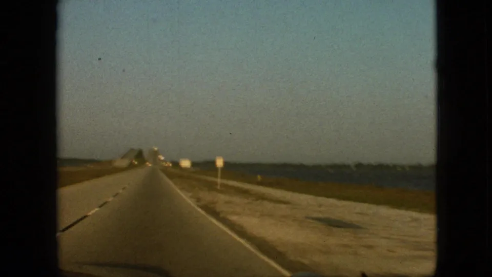 Moving-car view of a highway beside water near Branson, Missouri, with a raised roadway section ahead and low roadside signs along the shoulder.