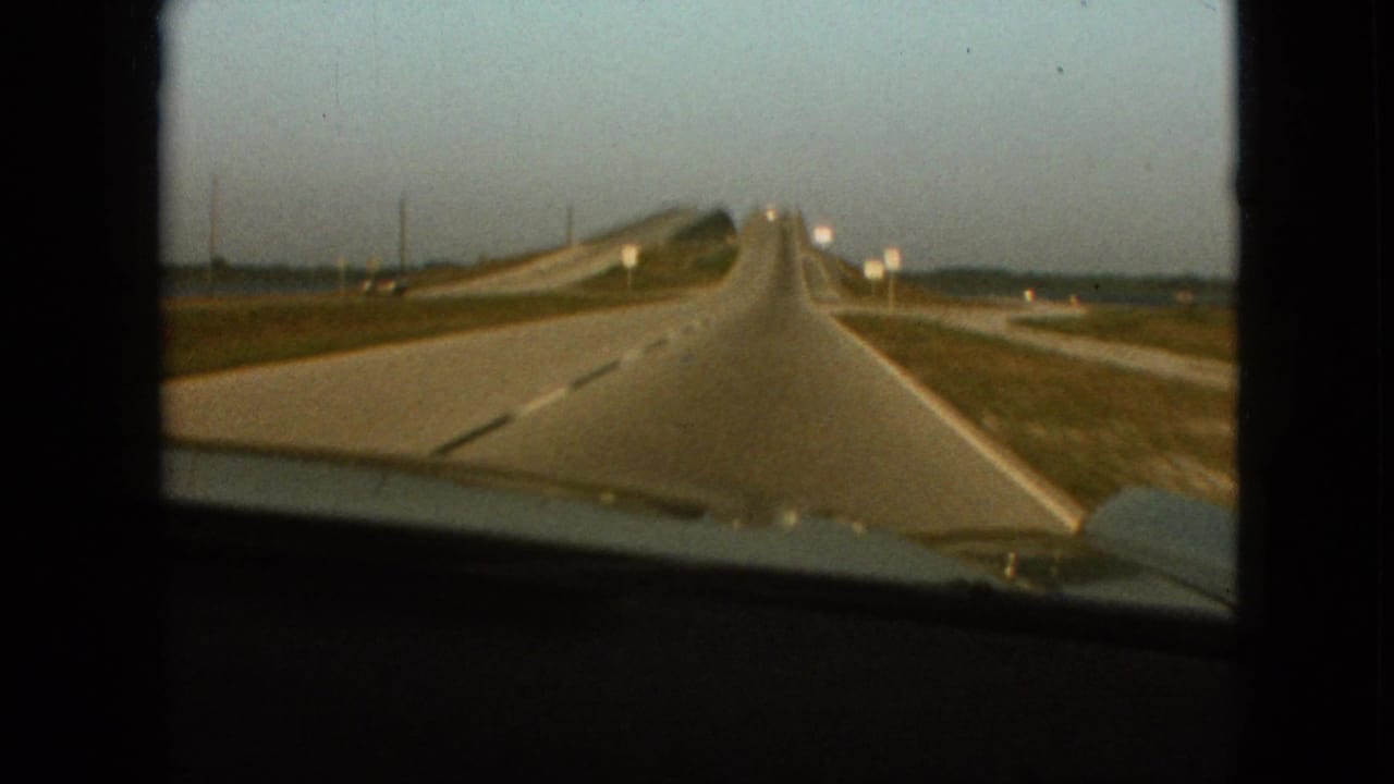 1968 8mm home-movie frame near Branson, Missouri, showing a forward windshield view of a straight highway, grassy shoulders, distant signs, open sky, and the car hood at the bottom edge.
