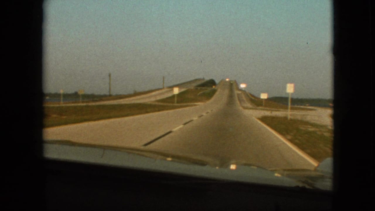 1968 8mm frame from a moving vehicle near Branson, Missouri, with the highway leading ahead, a raised or curving roadway on the left, sparse roadside signs, open sky, and the car hood visible below.