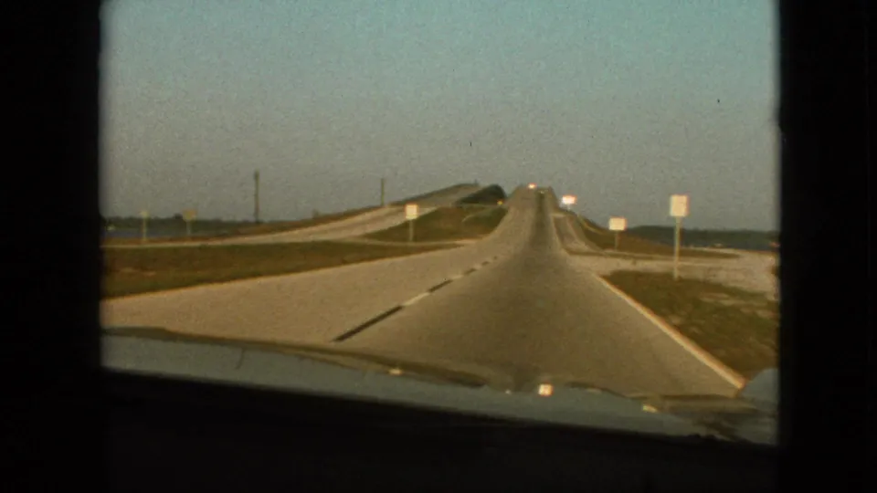 1968 8mm frame from a moving vehicle near Branson, Missouri, with the highway leading ahead, a raised or curving roadway on the left, sparse roadside signs, open sky, and the car hood visible below.