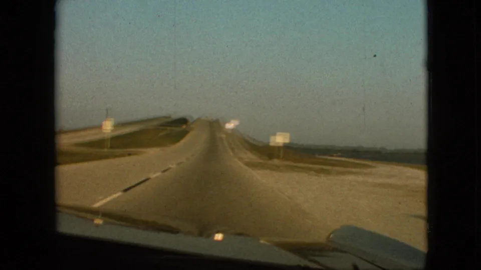 1968 8mm home-movie view near Branson, Missouri, showing the car approaching a broad highway area with signs, a light shoulder or side road, grassy banks, distant terrain, and windshield framing.
