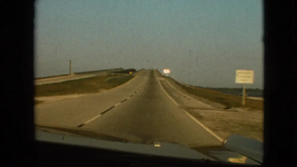 1968 8mm frame near Branson, Missouri, showing a straight highway continuing toward the horizon with dashed markings, roadside signs, low grassy landscape, open sky, and the car hood at the frame bottom.
