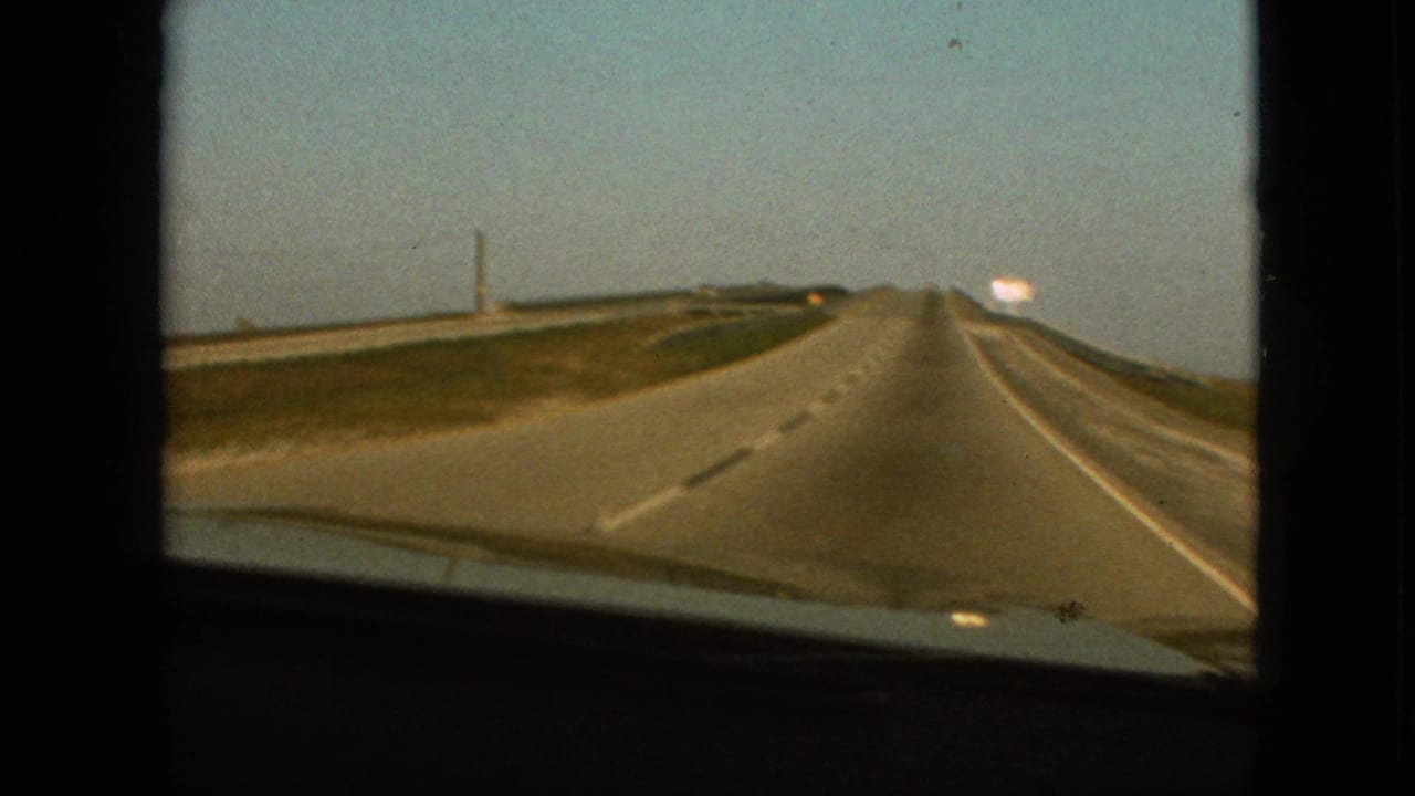 1968 8mm home-movie frame from the supplied Branson, Missouri clip showing a passenger's forward windshield view down a straight highway with grassy shoulders and a distant bright roadside sign.