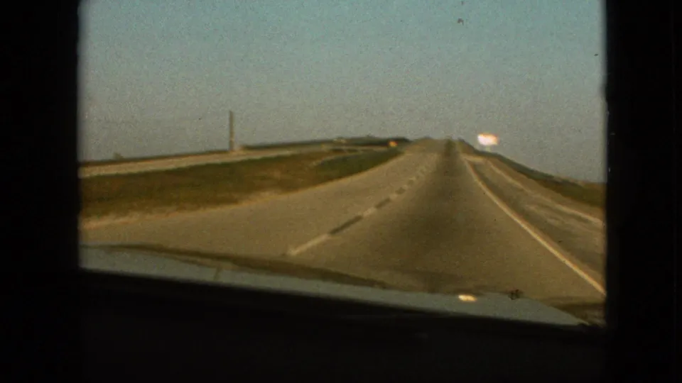 1968 8mm home-movie frame from the supplied Branson, Missouri clip showing a passenger's forward windshield view down a straight highway with grassy shoulders and a distant bright roadside sign.