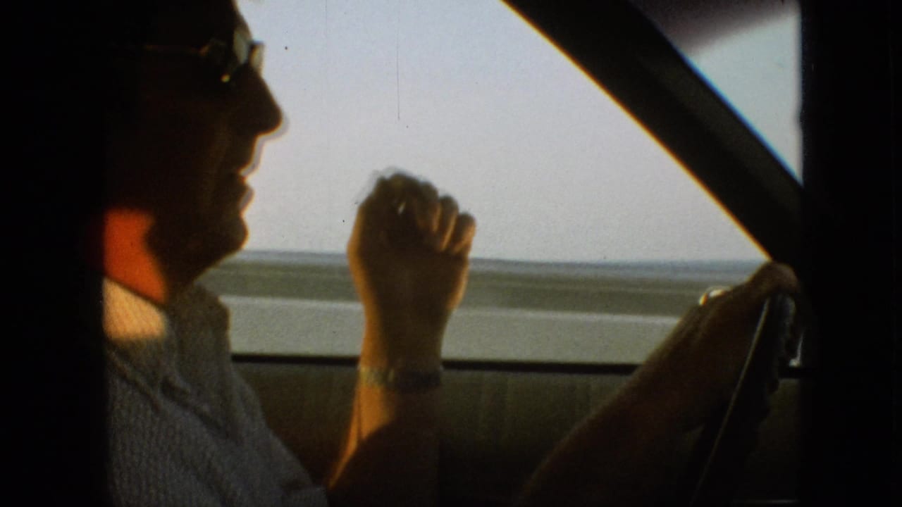 1968 8mm home-movie frame from the supplied Branson, Missouri clip showing a man in sunglasses and a light shirt driving, with his hand raised near his face and the road landscape visible through the side window.