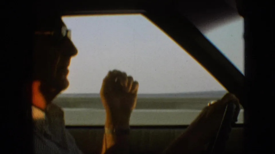 1968 8mm home-movie frame from the supplied Branson, Missouri clip showing the driver in profile inside the car, his hand near his face, with strong sunlight, the side window frame, and flat roadside scenery outside.