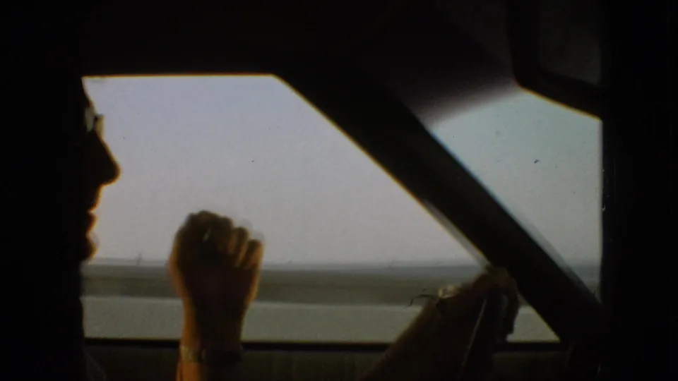 1968 8mm home-movie frame from the supplied Branson, Missouri clip showing a darker silhouetted profile of the driver, a hand near his face, the steering wheel, diagonal window frame, and low roadside horizon beyond the car window.