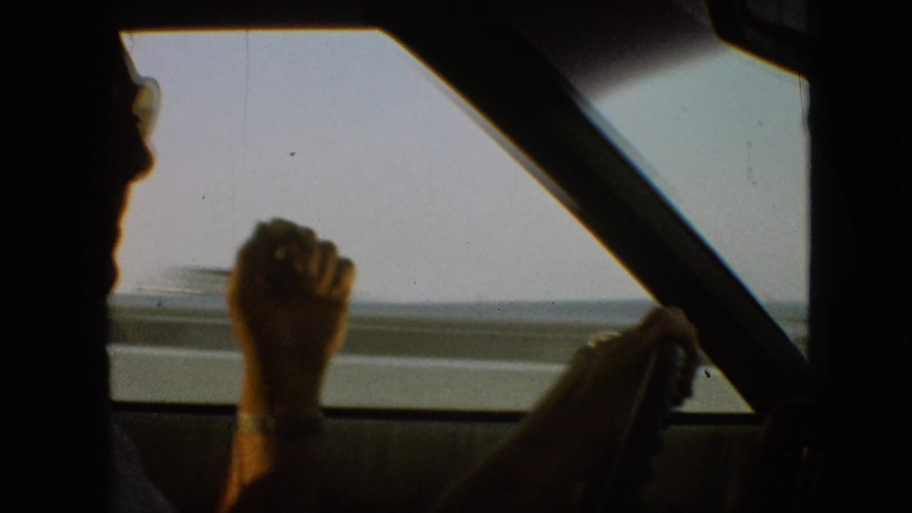 Passenger-seat view of an adult male driver in silhouette holding the steering wheel during a 1968 Missouri road trip, with bright water or open landscape outside the side window.