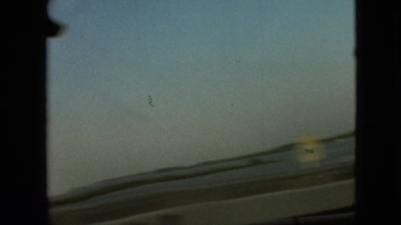 Forward windshield view of a highway bordered by water and low roadside structures.