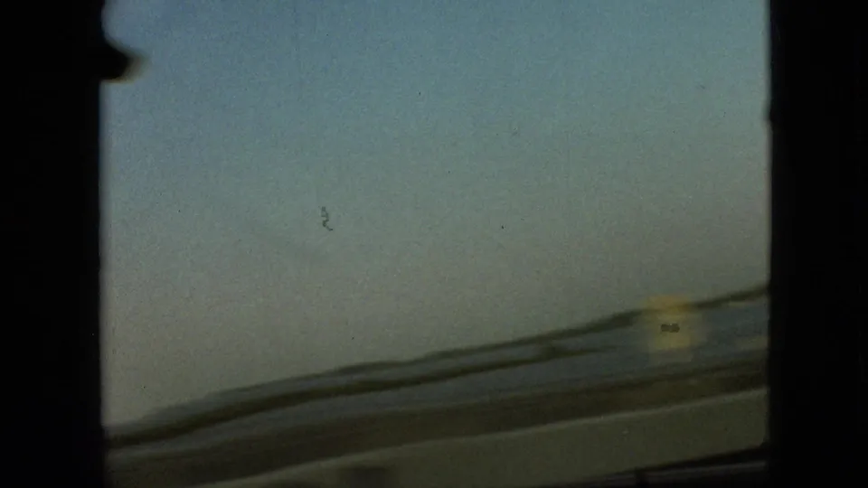 Forward view through a moving car windshield on a waterside highway in the supplied Branson, Missouri 1968 clip, with road edge and water visible under a pale sky.