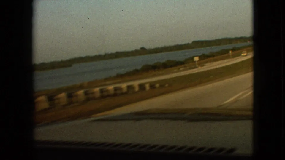 Moving-car view of a highway running alongside water near Branson, Missouri in 1968, with guardrails, shoreline, road signs, and a small vehicle ahead or approaching.