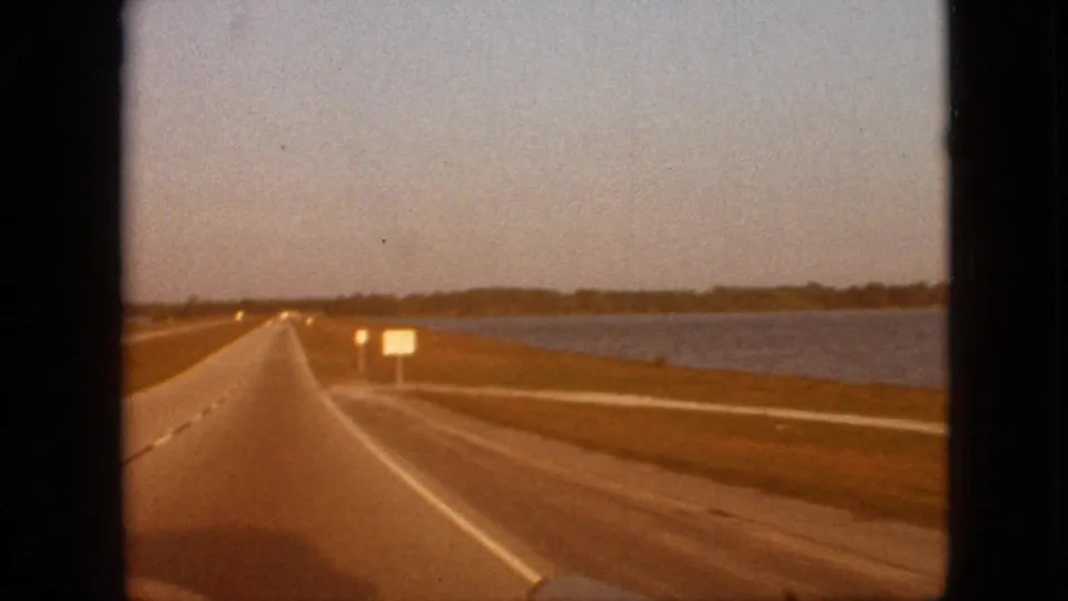 Forward passenger-car view down a straight waterside highway in the supplied 1968 Branson, Missouri footage, with lane markings, road signs, water, and distant tree line.