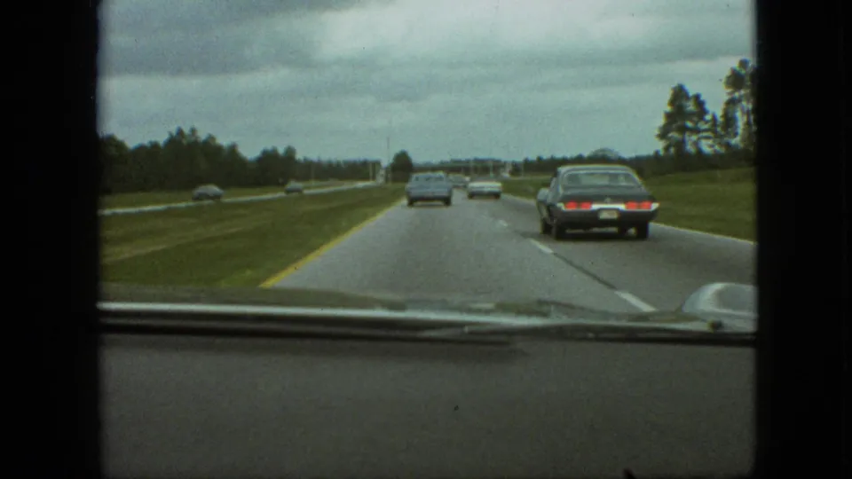 Forward-facing 8mm car-window view from the 1968 road-trip footage, showing highway traffic, grassy shoulders, trees, and an overcast sky.