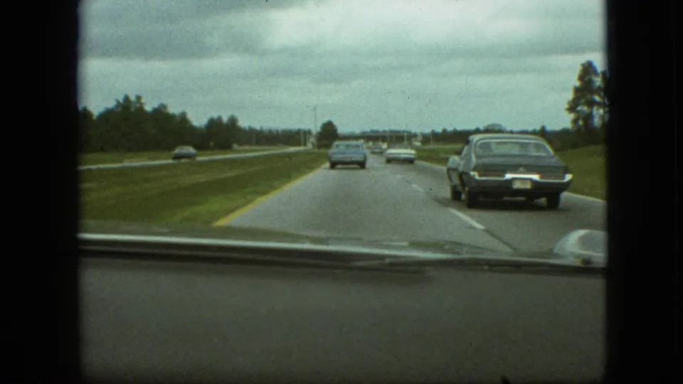 1968 8mm home-movie view from inside a moving vehicle, with a dark sedan passing on the right side of a divided highway under cloudy light.