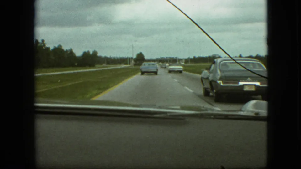 Late-1960s road-trip frame from inside a car, showing highway lanes, vehicles ahead, a dark sedan near the right edge, and visible film scratch over the scene.