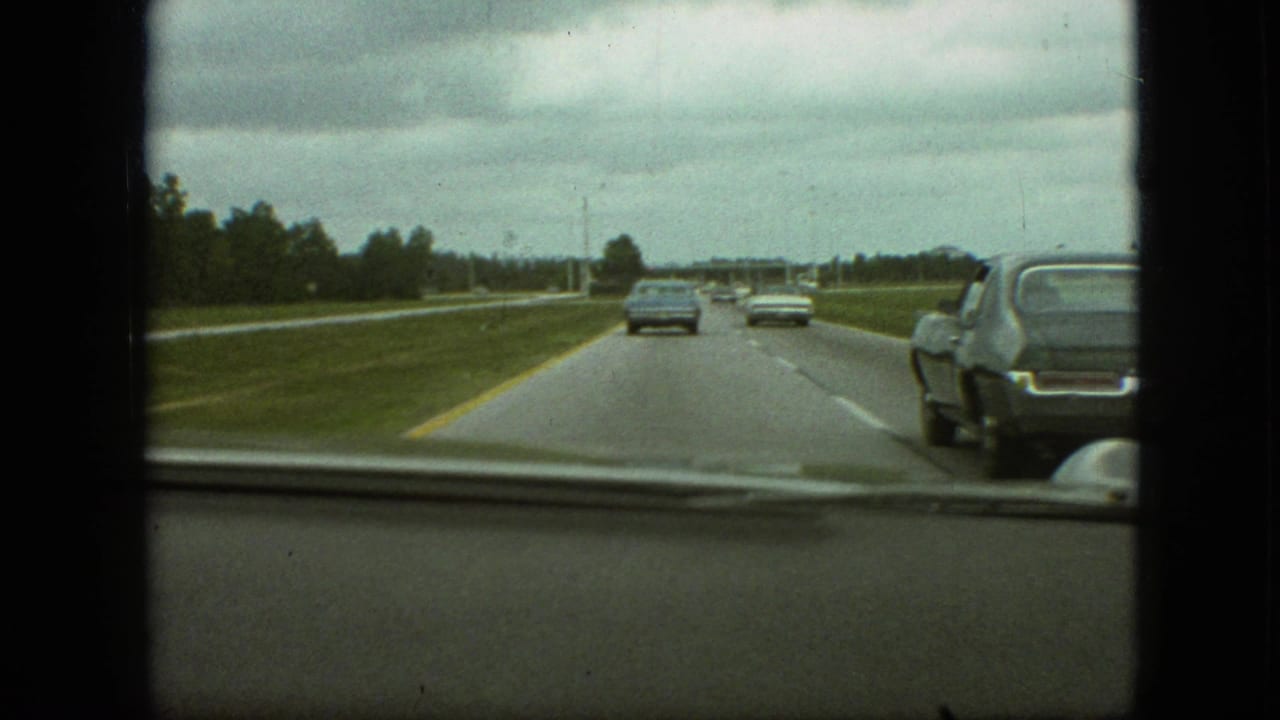 1968 Branson, Missouri home-movie view through a car windshield, showing a cloudy highway drive with several period automobiles and a dark sedan overtaking on the right.
