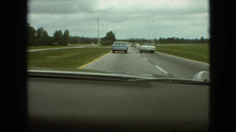 Passenger-view 8mm frame from the 1968 Branson, Missouri clip, showing period cars ahead on a damp-looking roadway with open grass and roadside trees.