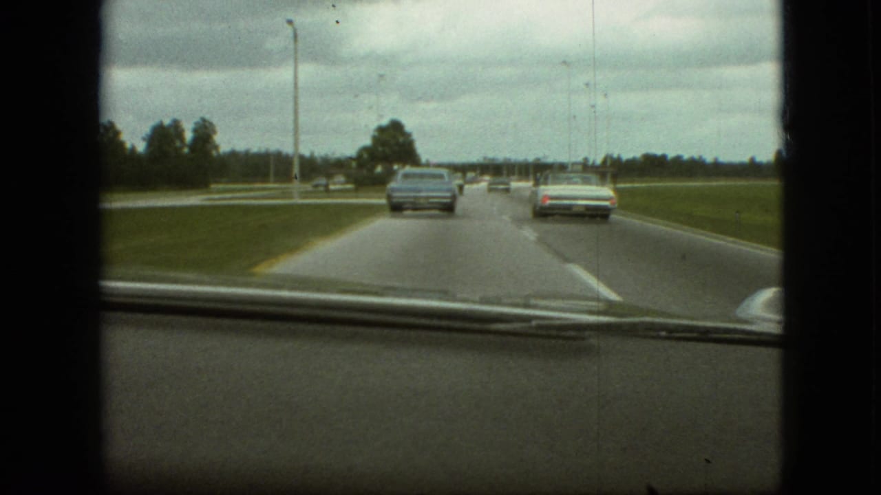 1968 Branson, Missouri 8mm dashboard view showing a light blue sedan and a pale car ahead on a broad roadway bordered by grass and trees.