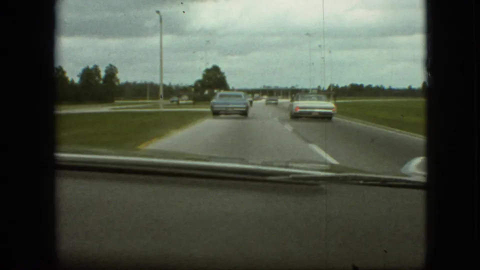 1968 Branson, Missouri 8mm dashboard view showing a light blue sedan and a pale car ahead on a broad roadway bordered by grass and trees.