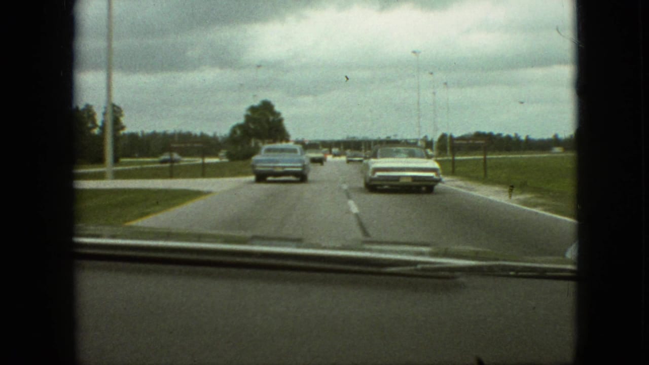 Classic cars drive ahead on a divided road with grass shoulders and tall light poles.