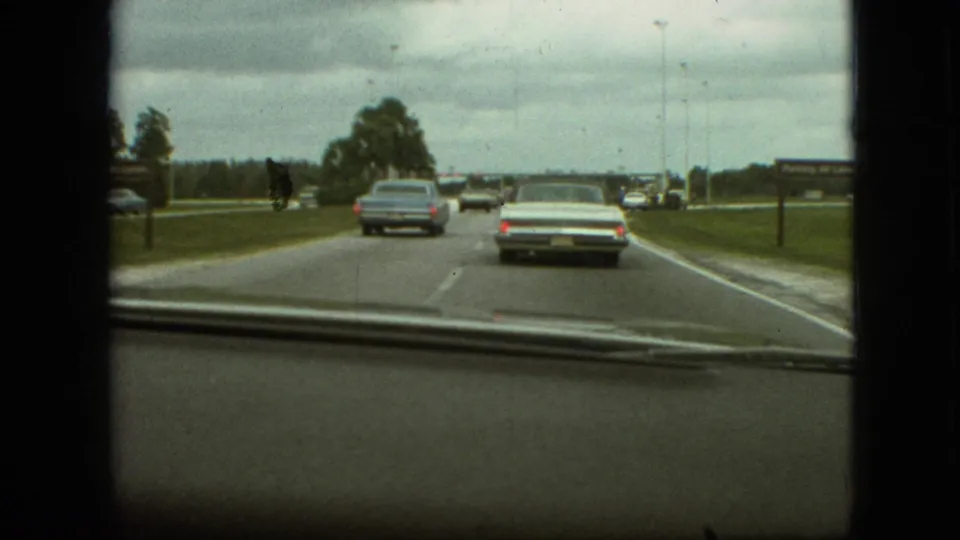 1968 Branson, Missouri dashboard shot showing a white convertible and blue sedan on a gently curving road with open grass and trees nearby.