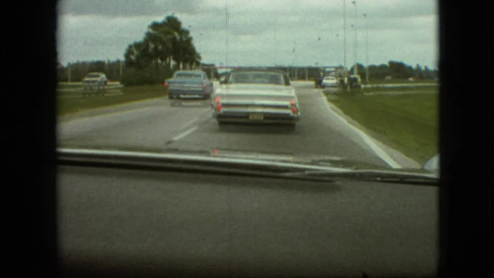 Dashboard view from 1968 Branson, Missouri, with a white convertible close ahead, a blue sedan farther forward, and grassy roadside areas on both sides.