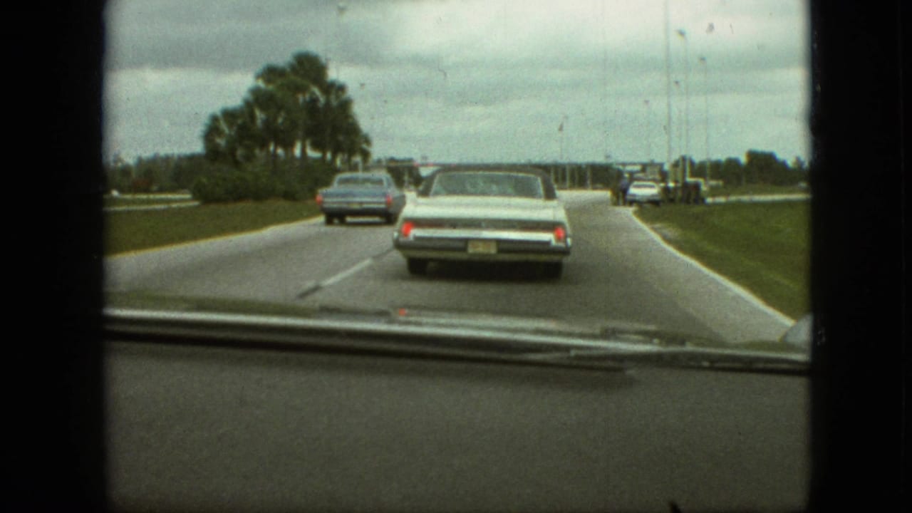 1968 home-movie windshield view from a moving car in the supplied Branson, Missouri clip, following pale and blue sedans on a broad road lined with palms.