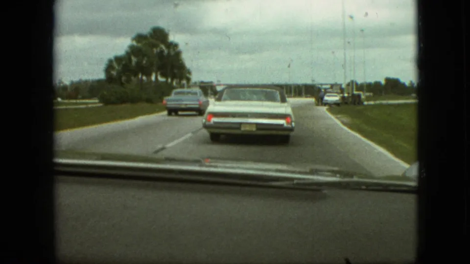 1968 home-movie windshield view from a moving car in the supplied Branson, Missouri clip, following pale and blue sedans on a broad road lined with palms.