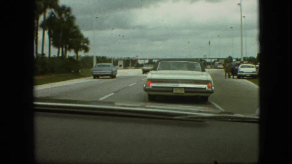 The home-movie camera follows a pale sedan on the road while a blue car passes left and several distant people appear near vehicles at the right shoulder.