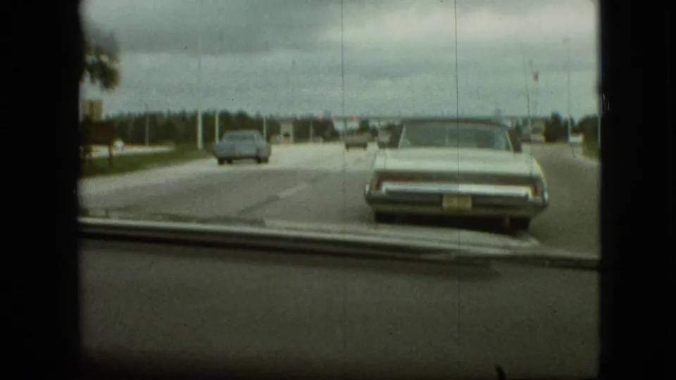 Late-1960s in-car road view from the supplied Branson, Missouri clip, showing a pale sedan ahead, a blue car farther left, and soft 8mm framing through the windshield.