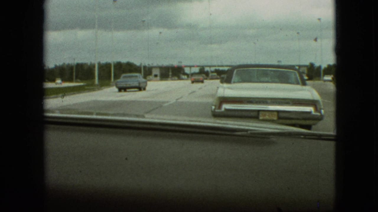 1968 Missouri home-movie windshield view from a moving car, showing a wide highway, classic automobiles, dashboard edge, and a distant toll plaza under an overcast sky.