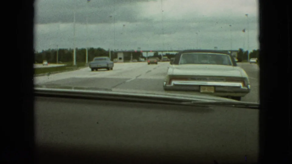 1968 Missouri home-movie windshield view from a moving car, showing a wide highway, classic automobiles, dashboard edge, and a distant toll plaza under an overcast sky.