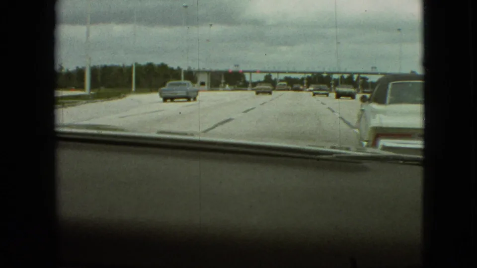 Windshield-framed 1968 Missouri highway footage shows several classic cars moving toward a toll plaza, with the car interior edge visible at the bottom of the frame.