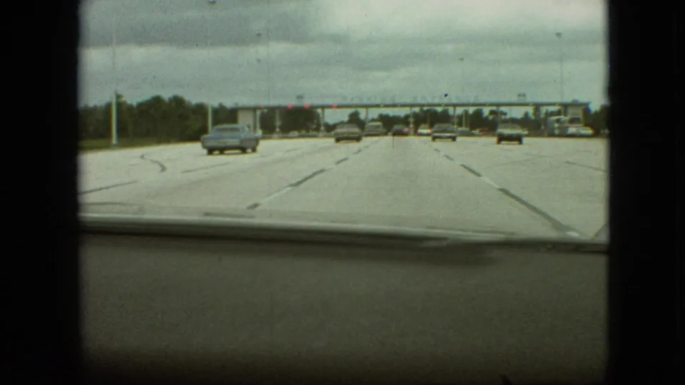Passenger-car windshield view on a 1968 Missouri highway, with multiple lanes, scattered classic automobiles, road markings, and a toll plaza visible ahead.