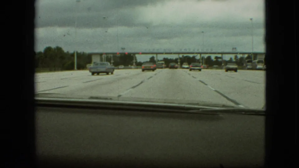 Late-1960s Missouri home-movie frame from inside a moving car, showing classic highway traffic, a dashboard edge, lane markings, and toll booths in the distance.